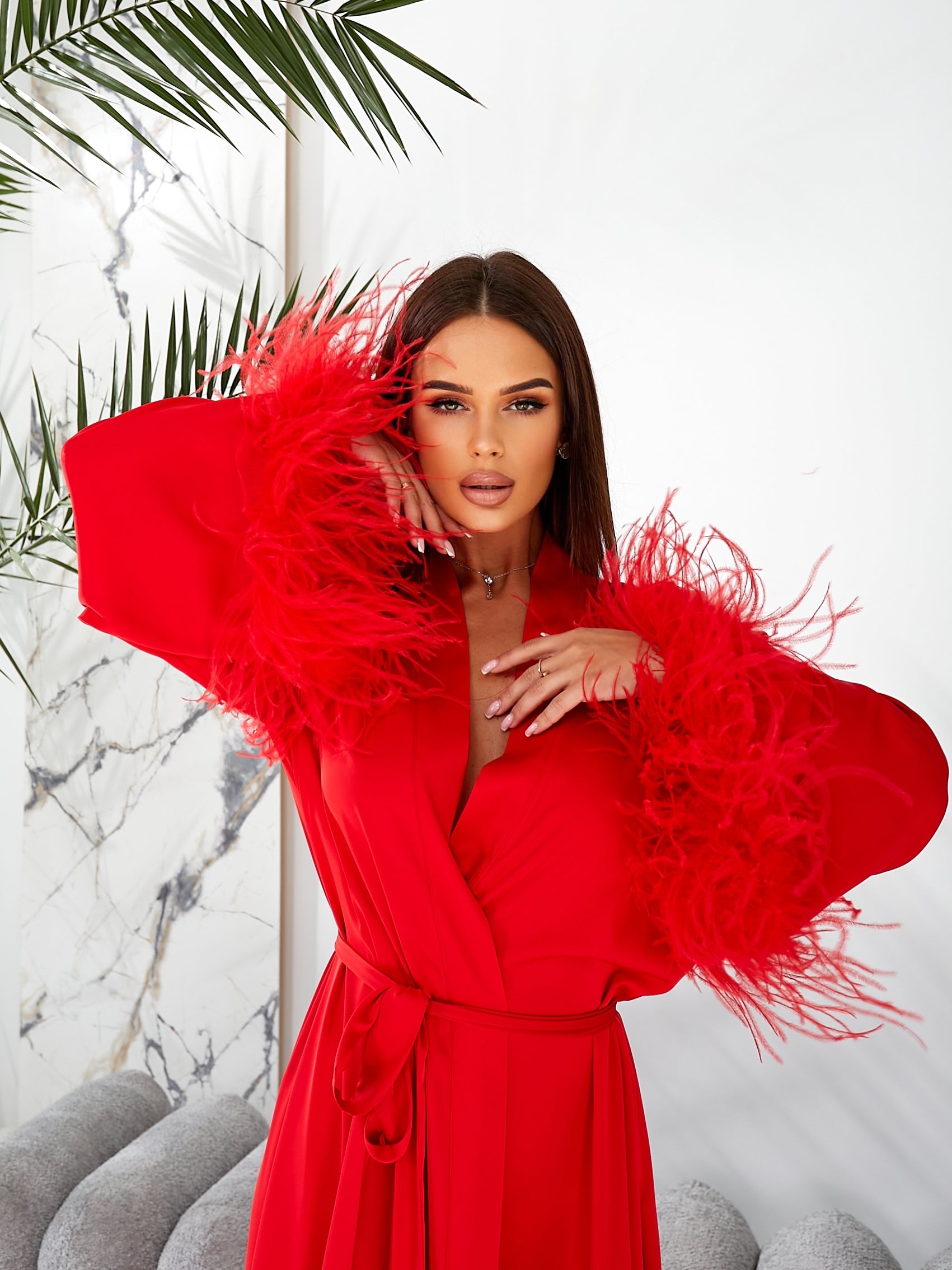 Boudoir robe bridal Woman wearing a red dress with feathered sleeves against a marble wall.