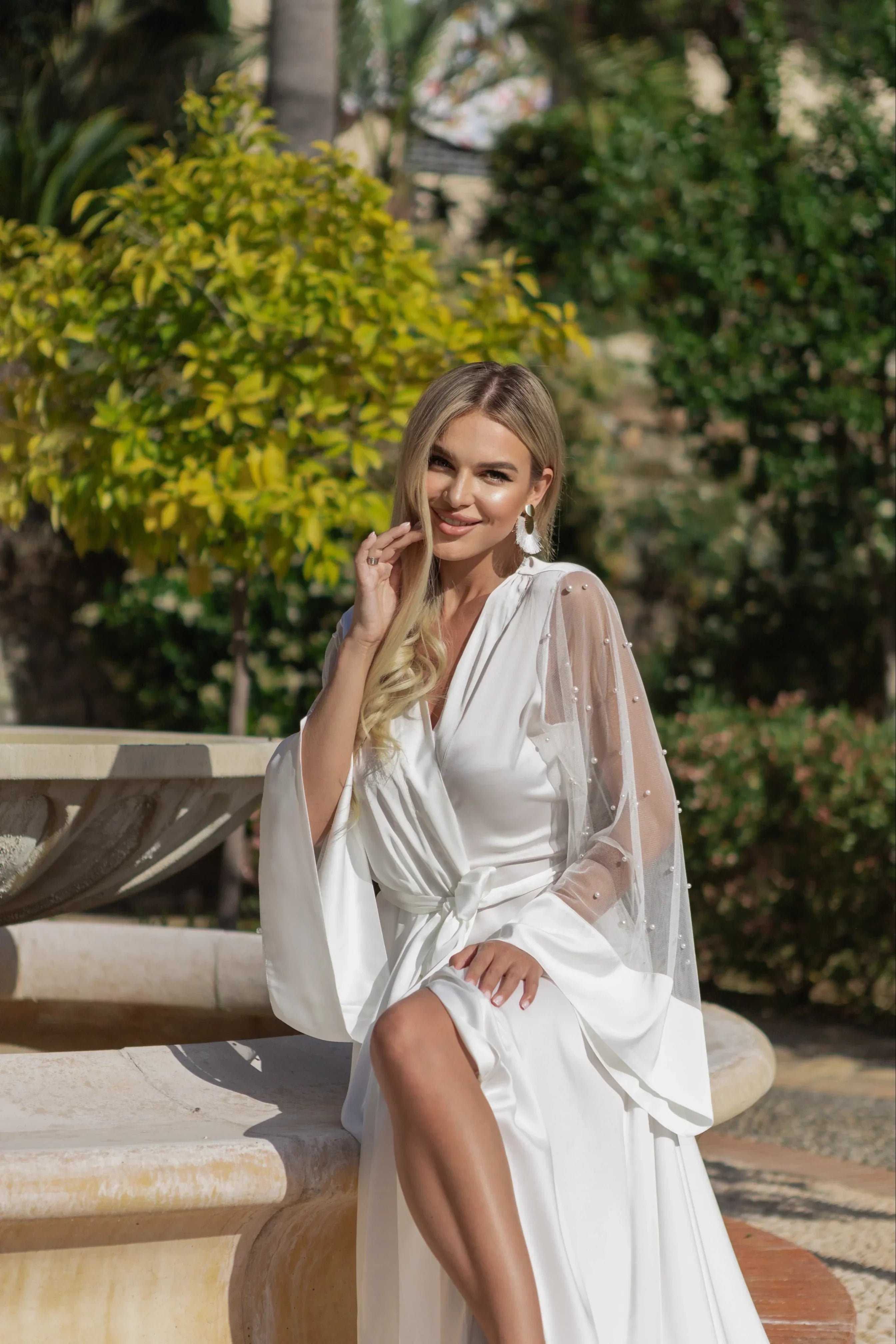 Bride wearing a white silk bridal robe with sheer pearl-detailed sleeves, sitting by a fountain outdoors.